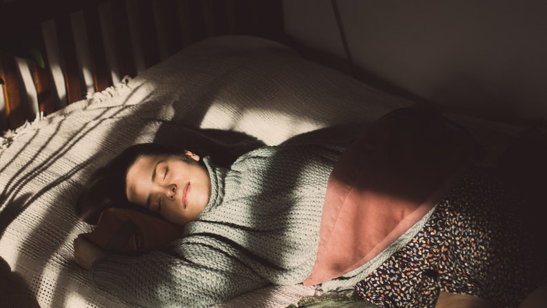 May include: A person sleeping in a bed with a white blanket and a pink pillow. The person is wearing a light green sweater and has dark hair.