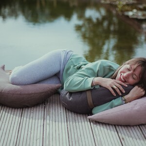 A woman with a joyful smile lounges on a wooden deck by a pond, hugging a dark gray WELLB meditation cushion. She rests on soft mauve and brown crescent-shaped pillows. A peaceful outdoor relaxation scene.