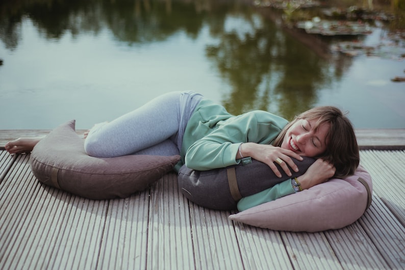 A woman with a joyful smile lounges on a wooden deck by a pond, hugging a dark gray WELLB meditation cushion. She rests on soft mauve and brown crescent-shaped pillows. A peaceful outdoor relaxation scene.