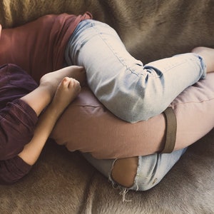A cozy scene of a mother and child cuddling on a soft fur-covered surface. The mother, wearing light blue jeans and a rust-colored top, supports her legs with a mauve WELLB crescent-shaped meditation cushion. The child, rests against her.