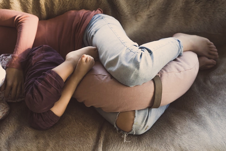 A cozy scene of a mother and child cuddling on a soft fur-covered surface. The mother, wearing light blue jeans and a rust-colored top, supports her legs with a mauve WELLB crescent-shaped meditation cushion. The child, rests against her.
