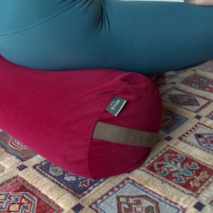 May include: A red meditation cushion with a brown strap and a green label that says "WELLO". The cushion is on a patterned rug.