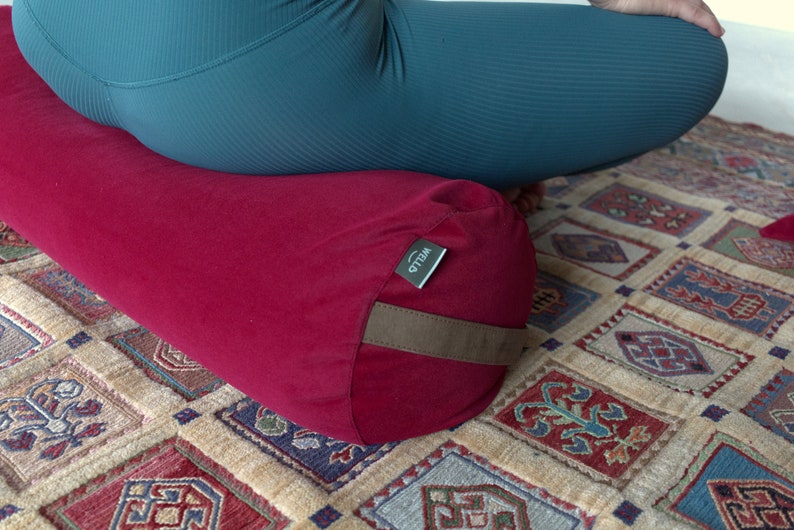 May include: A red meditation cushion with a brown strap and a green label that says "WELLO". The cushion is on a patterned rug.