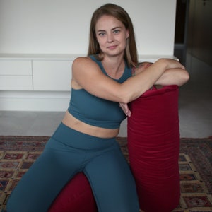 May include: A woman wearing teal blue leggings and a matching crop top is kneeling on a red yoga bolster. She is leaning on a larger red yoga bolster. The bolsters are on a patterned rug.