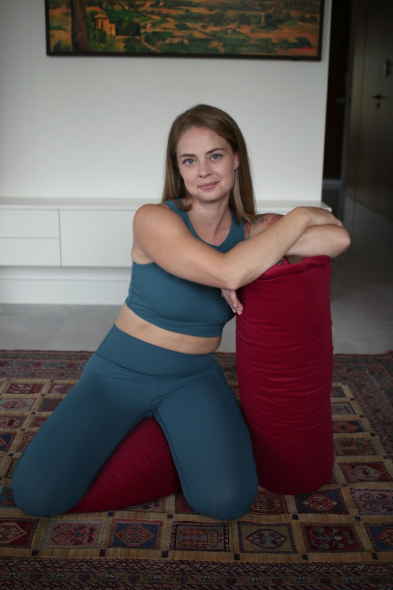May include: A woman wearing teal blue leggings and a matching crop top is kneeling on a red yoga bolster. She is leaning on a larger red yoga bolster. The bolsters are on a patterned rug.