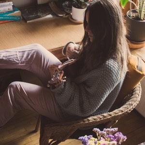 May include: A woman sits in a wicker chair with a yellow cushion, playing a ukulele. She is wearing a light blue sweater and pink pants. Her feet are resting on a wooden desk with books stacked on it. There are plants in pots on the windowsill behind her.