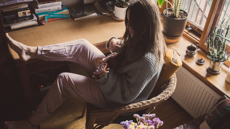 May include: A woman sits in a wicker chair with a yellow cushion, playing a ukulele. She is wearing a light blue sweater and pink pants. Her feet are resting on a wooden desk with books stacked on it. There are plants in pots on the windowsill behind her.