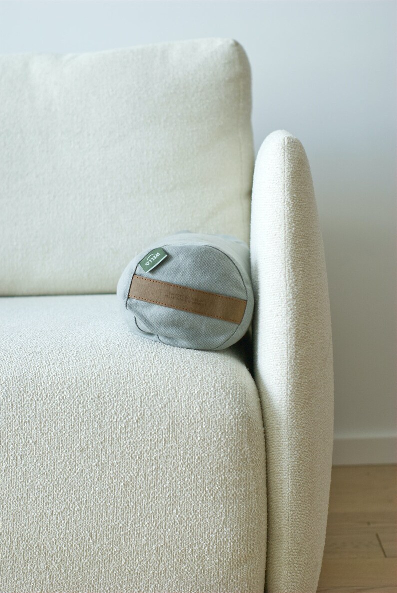 May include: A light gray cylindrical pillow with a brown leather strap and a green label that says "WILO" on a white couch.