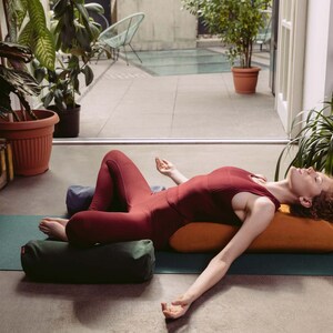 May include: A woman in a red tank top and black leggings is lying on a green yoga mat with two orange and green bolsters. She is practicing a restorative yoga pose with her arms outstretched and eyes closed.
