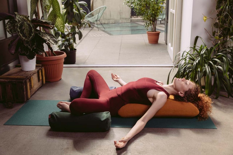 May include: A woman in a red tank top and black leggings is lying on a green yoga mat with two orange and green bolsters. She is practicing a restorative yoga pose with her arms outstretched and eyes closed.
