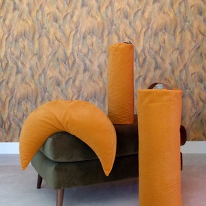A set of WELLB meditation cushions in mustard yellow, including a crescent-shaped pillow and two cylindrical bolsters. The background features textured, leaf-patterned wallpaper, creating a warm and stylish wellness space.
