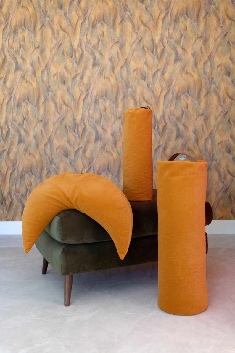 A set of WELLB meditation cushions in mustard yellow, including a crescent-shaped pillow and two cylindrical bolsters. The background features textured, leaf-patterned wallpaper, creating a warm and stylish wellness space.