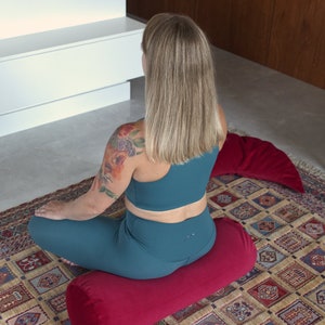 May include: A person sits on a red meditation cushion with a floral tattoo on their arm. The cushion is on a patterned rug.