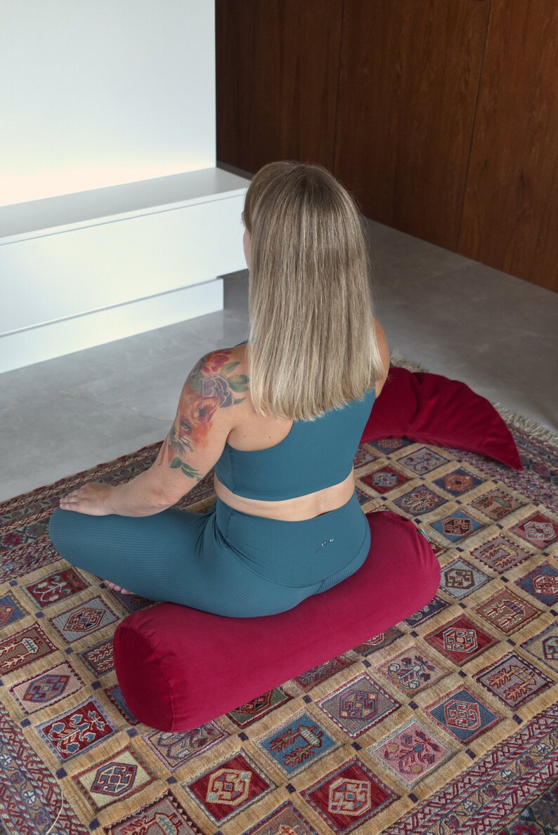 May include: A person sits on a red meditation cushion with a floral tattoo on their arm. The cushion is on a patterned rug.