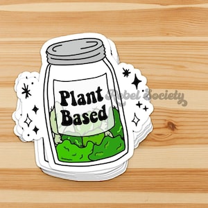 May include: A white sticker with a black outline of a mason jar filled with green leafy vegetables. The jar has a label that says "Plant Based" in black text. The sticker has a sparkly design with black stars and white sparkles.