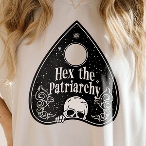 Może przedstawiać: Biała koszulka z czarnym nadrukiem w kształcie planszy Ouija z napisem 'Hex the Patriarchy'.