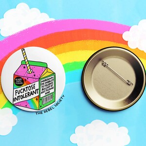 May include: A round button featuring a cartoon illustration of a juice carton with a straw. The juice carton is pink and green with a rainbow design. The text on the juice carton reads "Fucktose Intolerant". The button is white with a black border and the text "The Rebel Society" printed below the image.