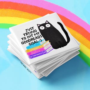 May include: A stack of white square magnets with a black cat illustration and text that reads "JUST TRYING TO HIT MY GOODREADS GOAL :)". The cat sits atop colorful book spines labeled "smut", "mystery", "fantasy", and "romance". A rainbow is in the background.