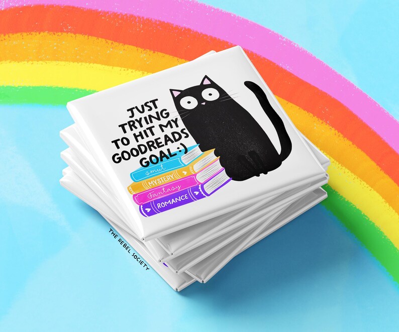 May include: A stack of white square magnets with a black cat illustration and text that reads "JUST TRYING TO HIT MY GOODREADS GOAL :)". The cat sits atop colorful book spines labeled "smut", "mystery", "fantasy", and "romance". A rainbow is in the background.