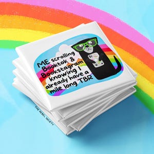 May include: A stack of white square magnets with a colorful illustration of a black otter wearing glasses and holding a phone. Text on the magnet reads "ME scrolling Booktok & Bookstagram knowing I already have a mile long TBR."