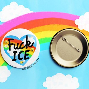 K&ouml;nnte beinhalten: Ein wei&szlig;er Button mit dem Text "Fuck ICE" in einem blauen Herzen mit Regenbogen und Smiley-Gesichtern. Der Button liegt neben einem Metall-Pinback auf blauem Hintergrund mit Regenbogen und Wolken.