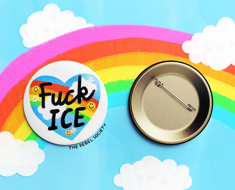 K&ouml;nnte beinhalten: Ein wei&szlig;er Button mit dem Text "Fuck ICE" in einem blauen Herzen mit Regenbogen und Smiley-Gesichtern. Der Button liegt neben einem Metall-Pinback auf blauem Hintergrund mit Regenbogen und Wolken.