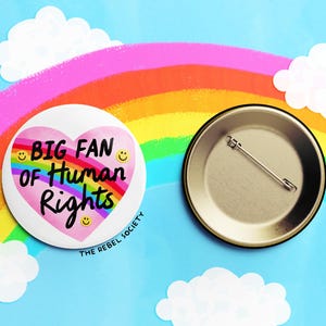 K&ouml;nnte beinhalten: Ein wei&szlig;er Ansteckbutton mit dem Text "BIG FAN of Human Rights" in einem rosa Herzen, umgeben von Smileys. Der Button befindet sich vor einem blauen Hintergrund mit einem Regenbogen und Wolken. Die R&uuml;ckseite des Buttons ist ebenfalls sichtbar.