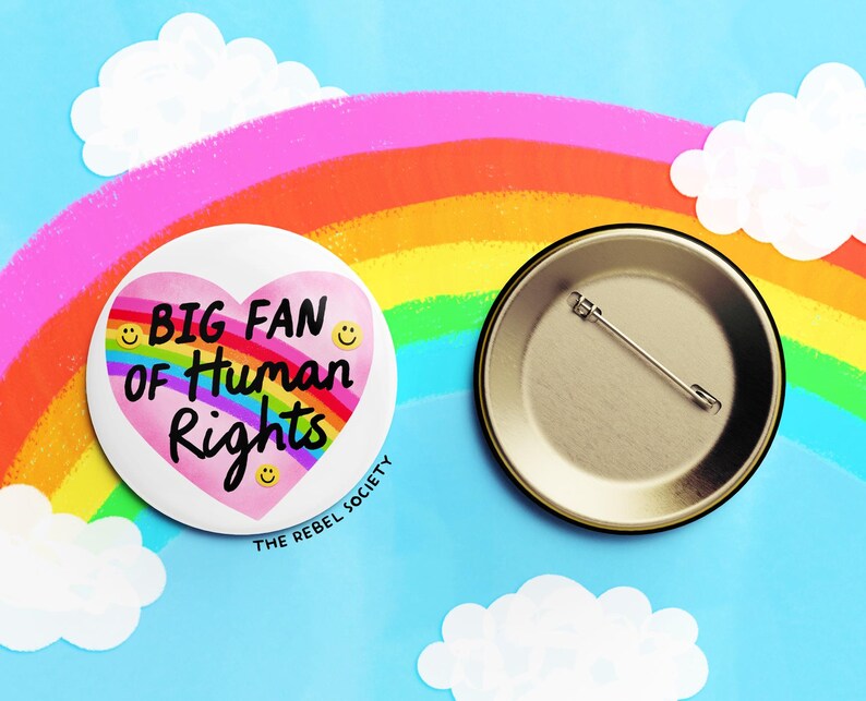 K&ouml;nnte beinhalten: Ein wei&szlig;er Ansteckbutton mit dem Text "BIG FAN of Human Rights" in einem rosa Herzen, umgeben von Smileys. Der Button befindet sich vor einem blauen Hintergrund mit einem Regenbogen und Wolken. Die R&uuml;ckseite des Buttons ist ebenfalls sichtbar.