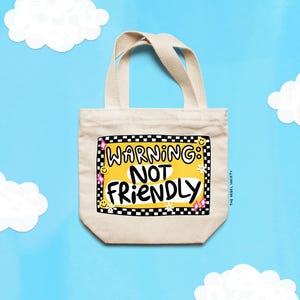 May include: A beige canvas tote bag with two handles featuring a central graphic with a black and white checkered border. The design includes the text WARNING: NOT FRIENDLY in black letters on a yellow background, accented with small smiley faces, flowers, and pink stars.