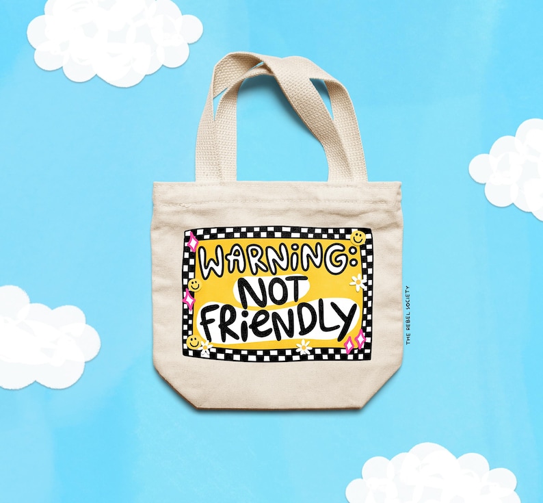 May include: A beige canvas tote bag with two handles featuring a central graphic with a black and white checkered border. The design includes the text WARNING: NOT FRIENDLY in black letters on a yellow background, accented with small smiley faces, flowers, and pink stars.