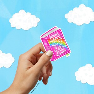 Puede incluir: Una pegatina rosa con forma de libro con el texto "BOOKS BECAUSE COPING WITH REALITY IS NOT AN OPTION AT THE MOMENT" y un dise&ntilde;o de arco&iacute;ris. La pegatina se sostiene sobre un fondo de cielo azul con nubes blancas.
