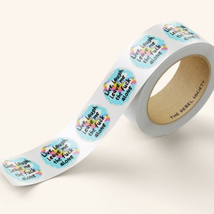May include: A roll of stickers with the text "Live, Laugh, Leave me the fuck alone" in a white cloud with a rainbow arc. The text is in black and the background is blue. The stickers are on a white roll with the text "THE REBEL SOCIETY" printed on the roll.
