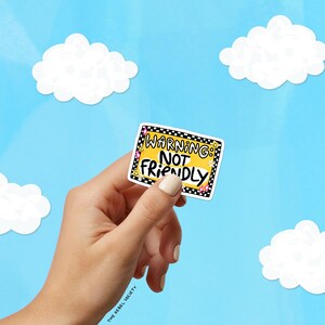 May include: A rectangular sticker with a yellow background and a black and white checkered border. The text reads WARNING: NOT FRIENDLY in bold black letters. The design includes small yellow smiley faces and pink star accents on the corners.