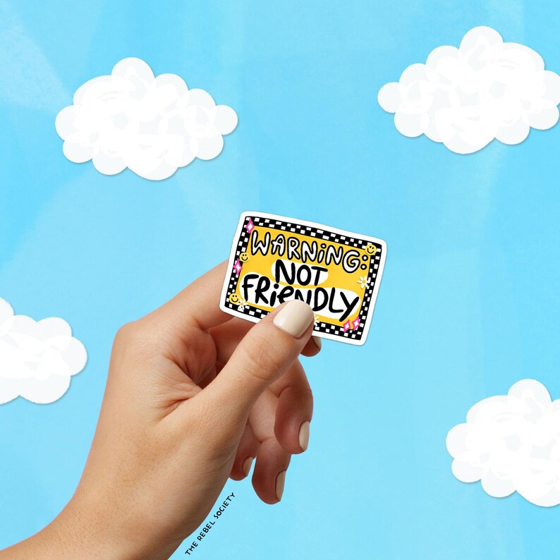 May include: A rectangular sticker with a yellow background and a black and white checkered border. The text reads WARNING: NOT FRIENDLY in bold black letters. The design includes small yellow smiley faces and pink star accents on the corners.