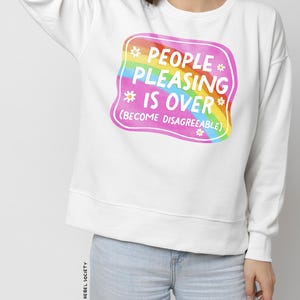 Puede incluir: Sudadera blanca con un gr&aacute;fico cuadrado rosa con un arco&iacute;ris y el texto "People Pleasing Is Over (Become Disagreeable)".