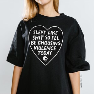 May include: Black t-shirt with a white heart-shaped graphic. The graphic includes the text "SLEPT LIKE SHIT SO I'LL BE CHOOSING VIOLENCE TODAY" and a small skull. The shirt is a casual style.