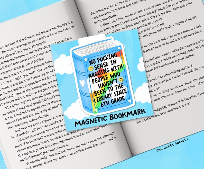 May include: A magnetic bookmark with a blue book design and the text "NO FUCKING SENSE IN ARGUING WITH PEOPLE WHO HAVEN'T BEEN TO THE LIBRARY SINCE 6TH GRADE." The bookmark is on a book.