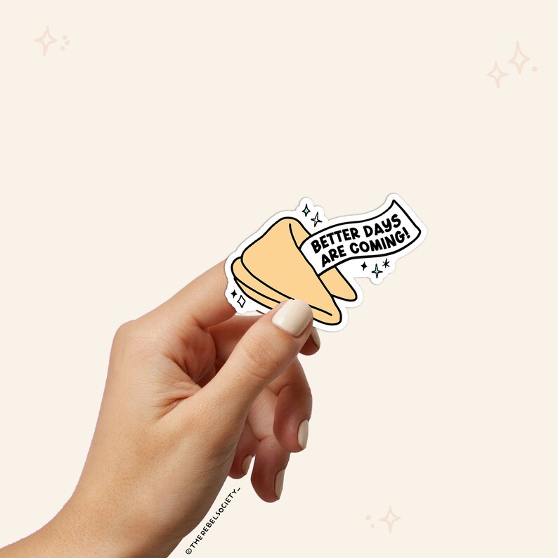 K&ouml;nnte beinhalten: Ein gelber Gl&uuml;ckskeks-Aufkleber mit dem Text "Better Days Are Coming!" in schwarzer Schrift. Der Aufkleber wird in einer Hand vor einem hellbeigen Hintergrund gehalten.
