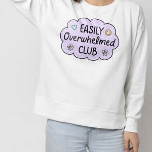 May include: White sweatshirt with a purple cloud graphic that says "EASILY Overwhelmed CLUB" with a heart, smiley face, and flowers.