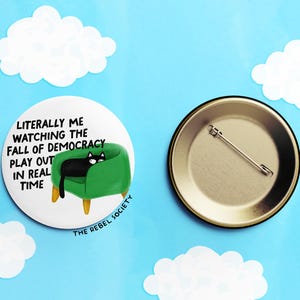 May include: A round button with a black cat lounging on a green chair, with the text "Literally me watching the fall of democracy play out in real time." The back of the button is gold with a pin.