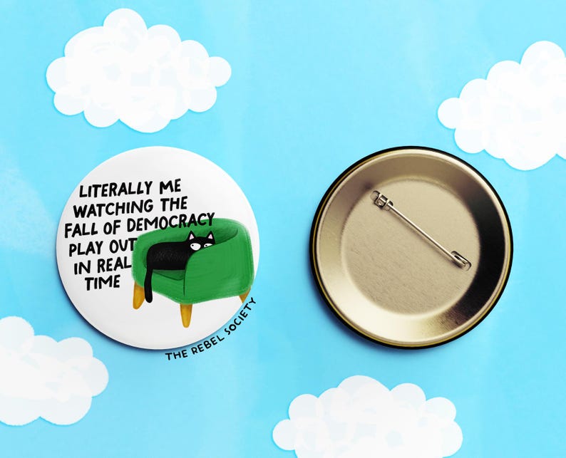 May include: A round button with a black cat lounging on a green chair, with the text "Literally me watching the fall of democracy play out in real time." The back of the button is gold with a pin.