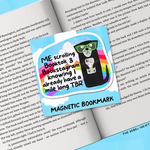 May include: A magnetic bookmark featuring a cartoon otter wearing green glasses and holding a phone. The bookmark has text that reads "ME scrolling Booktok & Bookstagram knowing I already have a mile long TBR". The bookmark is on a book.