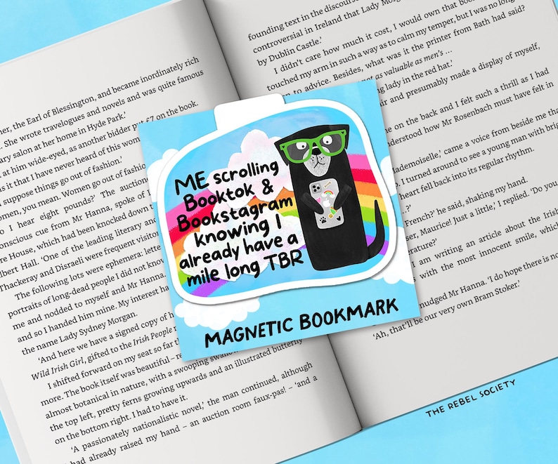 May include: A magnetic bookmark featuring a cartoon otter wearing green glasses and holding a phone. The bookmark has text that reads "ME scrolling Booktok & Bookstagram knowing I already have a mile long TBR". The bookmark is on a book.