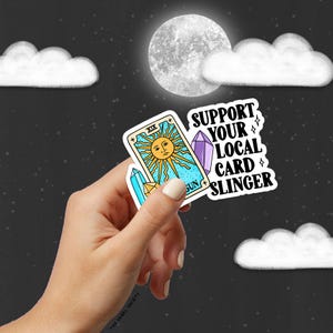 May include: A sticker featuring a tarot card design and the text "Support Your Local Card Slinger". The tarot card is the Sun card, depicted as a yellow sun with a face and rays. The card is surrounded by a blue and white border. The sticker is against a black background.
