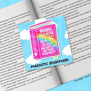 Puede incluir: Un marcap&aacute;ginas magn&eacute;tico con una ilustraci&oacute;n de libro rosa y el texto "BOOKS BECAUSE COPING WITH REALITY IS NOT AN OPTION AT THE MOMENT". Tiene un arco&iacute;ris y un coraz&oacute;n, sobre un cielo azul con nubes. Las palabras "MAGNETIC BOOKMARK" est&aacute;n en la parte inferior.