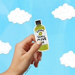 May include: A green sticker in the shape of an olive oil bottle with the words "EXTRA Slutty OLIVE Oil" on a white label. The sticker is held by a hand against a blue sky background with white cloud cutouts.