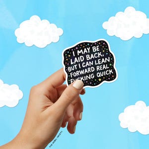 Op de afbeelding: Een zwarte sticker met de tekst "I MAY BE LAID BACK, BUT I CAN LEAN FORWARD REAL FUCKING QUICK" in het wit. De sticker wordt door een hand vastgehouden tegen een blauwe luchtachtergrond met witte wolken. Het logo van The Rebel Society staat linksonder.