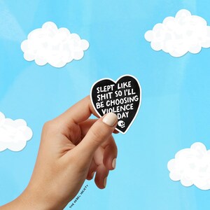 May include: A black heart-shaped sticker with white text that reads "SLEPT LIKE SHIT SO I'LL BE CHOOSING VIOLENCE DAY" and a skull graphic. The sticker is held against a blue sky background with white cloud cutouts.