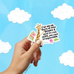 May include: A sticker featuring a giraffe with shopping bags from a "Local Book Shop" and the text "Y'all see ME Buying all these BOOKS & think I'm rich. NOPE, just super irresponsible." The sticker is held against a blue sky with white clouds.