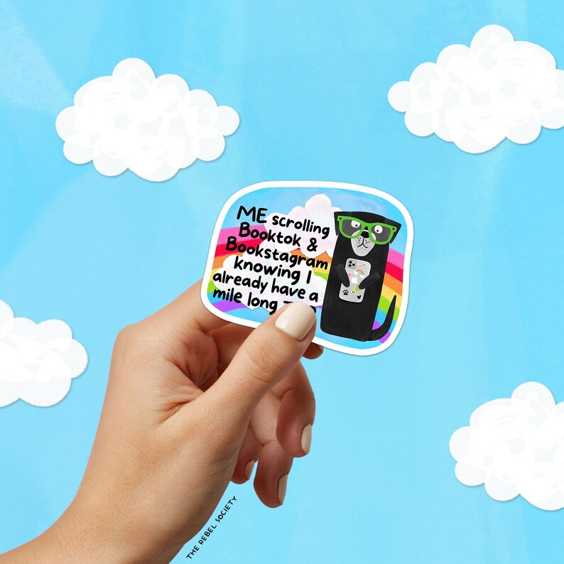 May include: A sticker featuring a black dog wearing green glasses and holding a phone. The sticker has text that reads "ME scrolling Booktok & Bookstagram knowing I already have a mile long." The background is blue with white clouds.